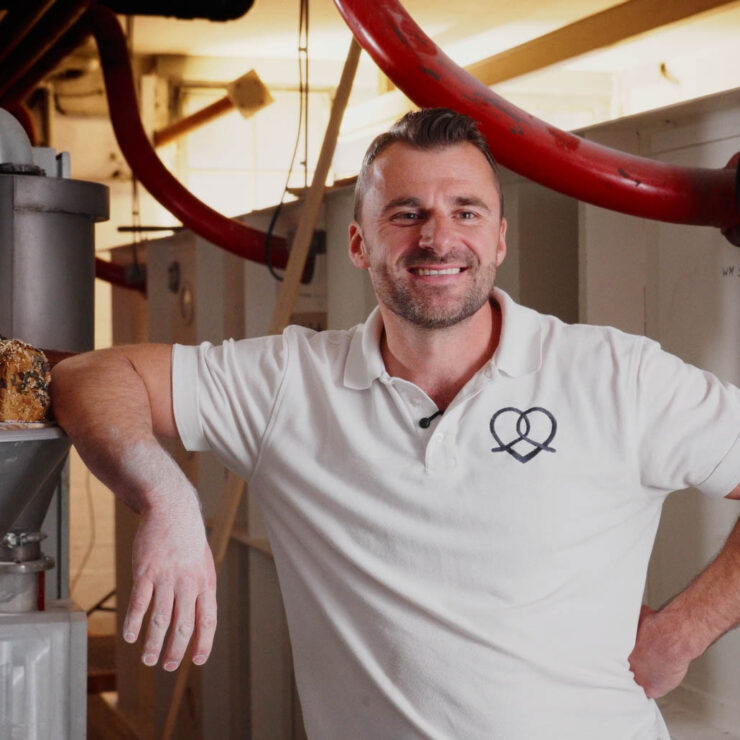 Ein Bäcker steht neben einer modernen Mühle. Es sind Brote zu sehen. Der Bäcker grinst in die Kamera.