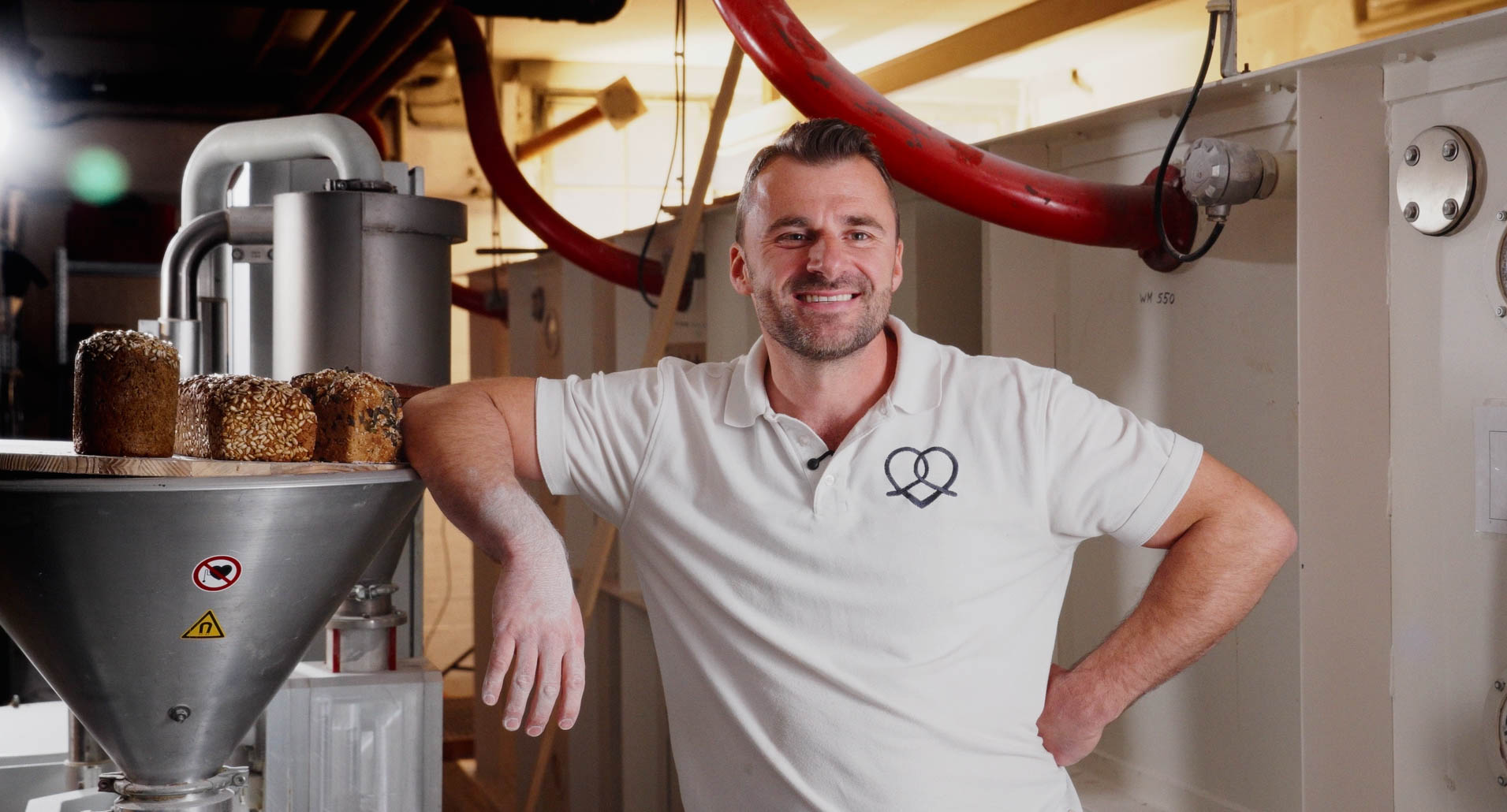 Ein Bäcker steht neben einer modernen Mühle. Es sind Brote zu sehen. Der Bäcker grinst in die Kamera.