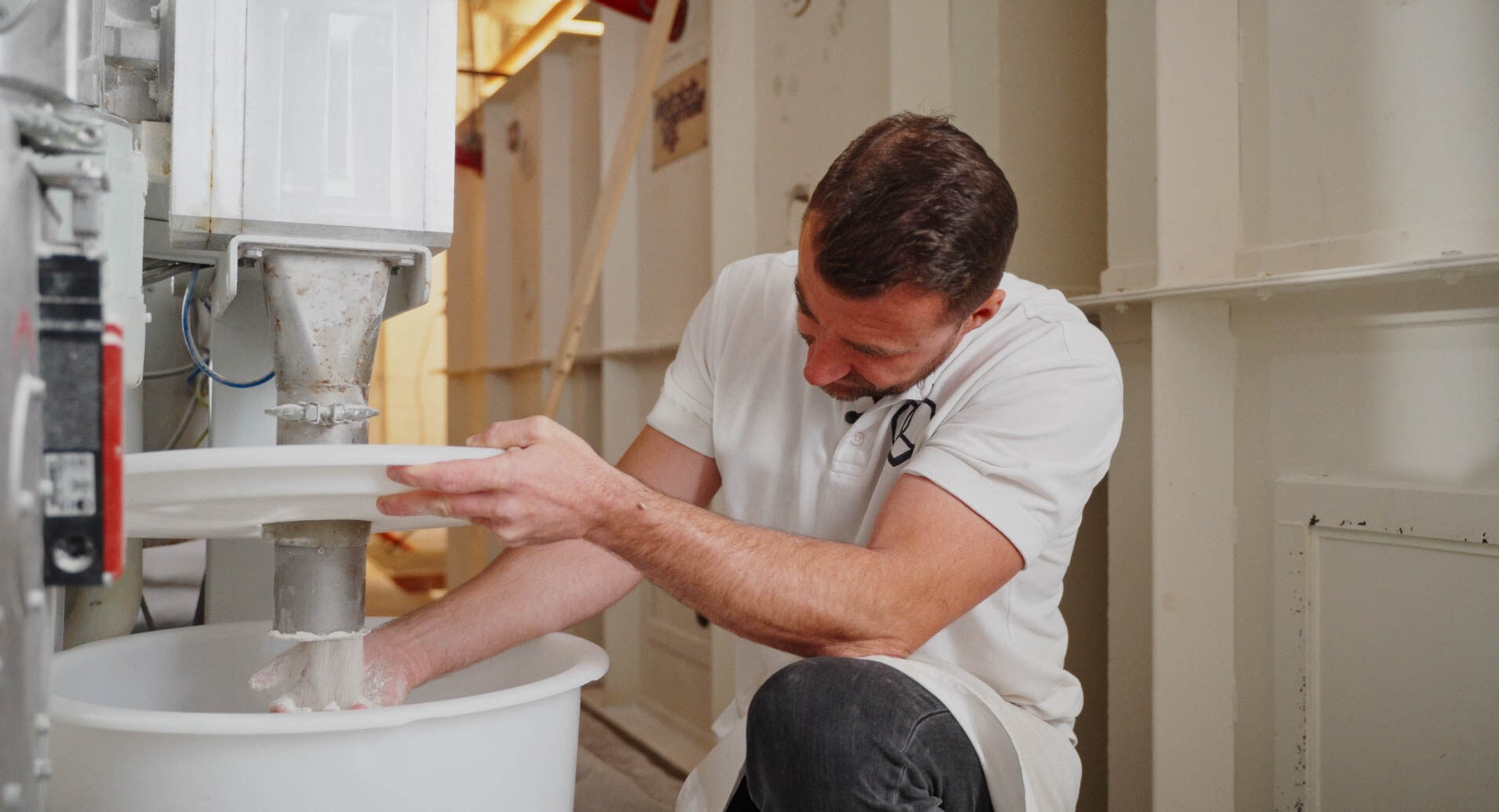 Ein Bäcker steht neben einer modernen Mühle und überprüft die Qualität des Mehls.