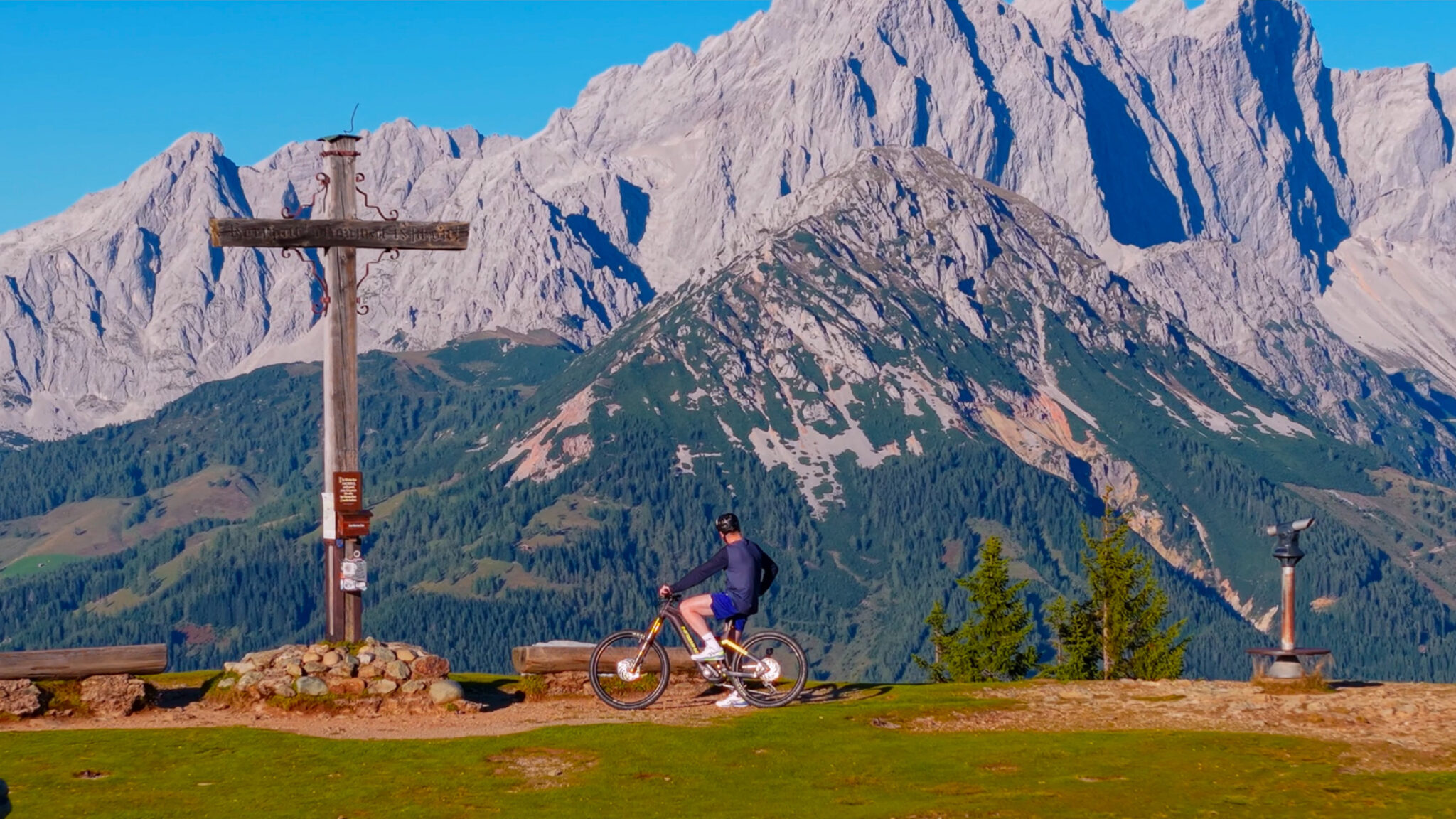 Spektakuläre Outdoor-Produktion von 4kFactory für ein Werbevideo für Hotels: Ein Mountainbiker am Gipfelkreuz vor dem massiven Bergpanorama in Flachau während des Dips & Drops Events.