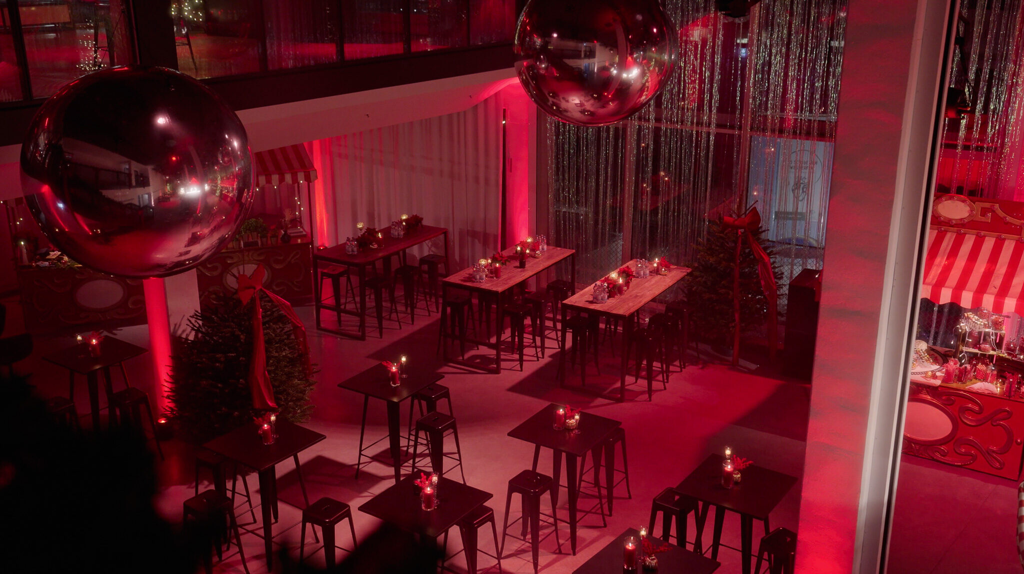 Professionelles Set-Design und Event-Beleuchtung von 4kFactory: Die leere Event-Fläche im Werk 3 München vor der Avantgard Weihnachtsfeier. Symmetrisch angeordnete Stehtische, festlich geschmückte Marktstände mit rot-weiß gestreiften Dächern und große Discokugeln unter einer Traversen-Konstruktion werden durch atmosphärisches rotes Licht für die Videoproduktion perfekt in Szene gesetzt.