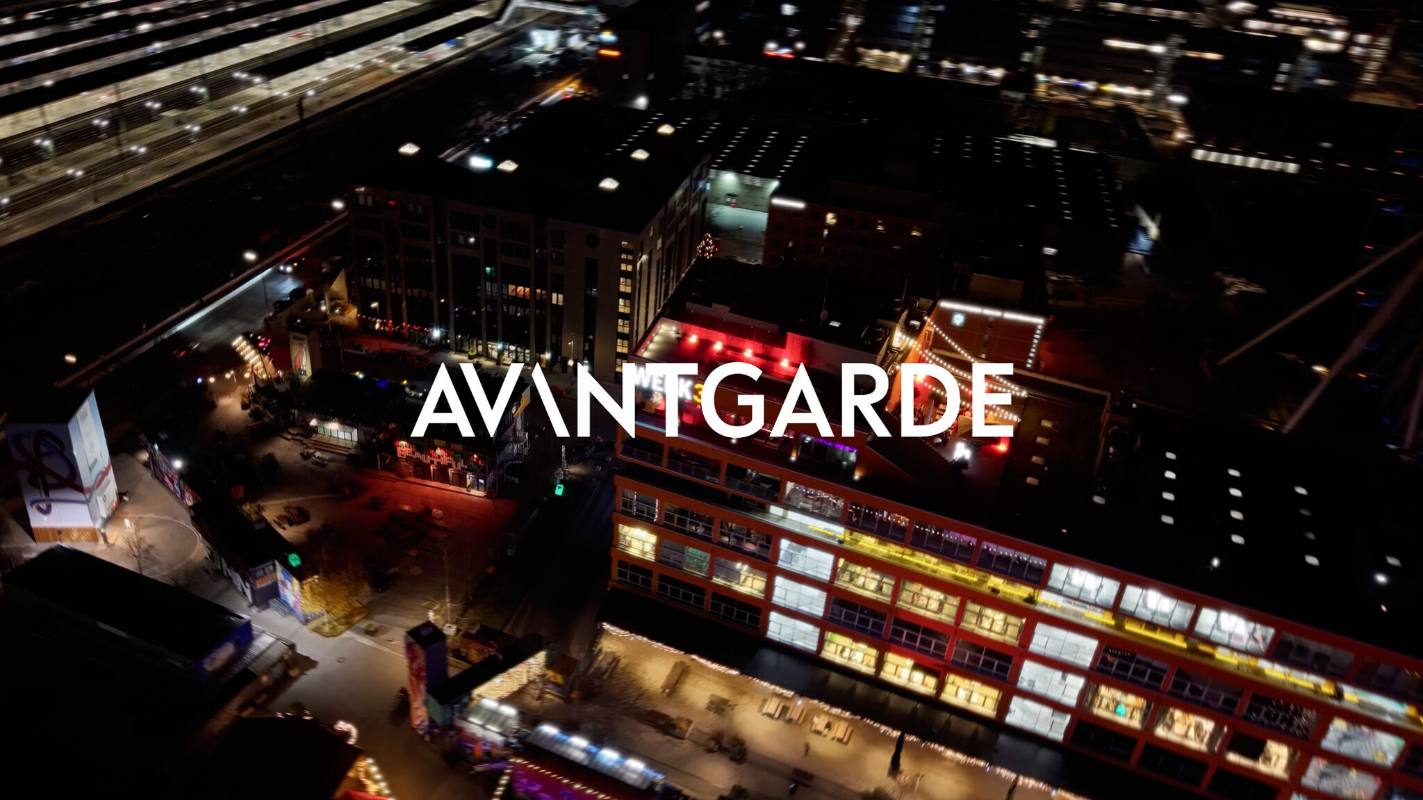 Drone-Shot und Videoproduktion der 4kFactory: Nächtliche Luftaufnahme vom Werk 3 in München während der Avantgard Weihnachtsfeier. Das beleuchtete Gebäude und das umliegende Werksviertel sind mit dem "AVANTGARDE"-Logo überlagert, was die cinematische Event-Berichterstattung unterstreicht.