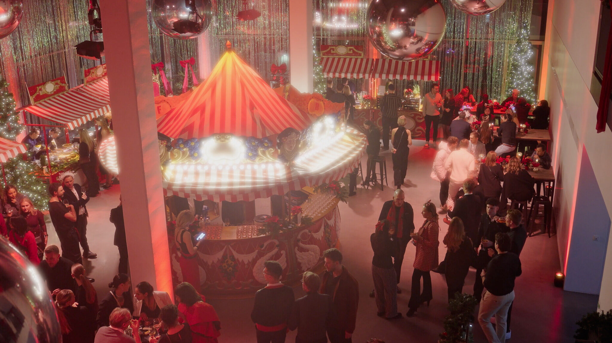 Blick von oben auf die Avantgard Weihnachtsfeier im Werk 3, München. Im Zentrum steht eine kreisförmige Bar in Form eines nostalgischen Jahrmarkts-Karussells mit rot-weiß gestreiftem Dach. Zahlreiche Gäste verteilen sich im festlich beleuchteten Raum, flankiert von Marktständen und glitzernden Vorhängen. Große, spiegelnde Discokugeln hängen von der Decke und reflektieren das warme Licht.