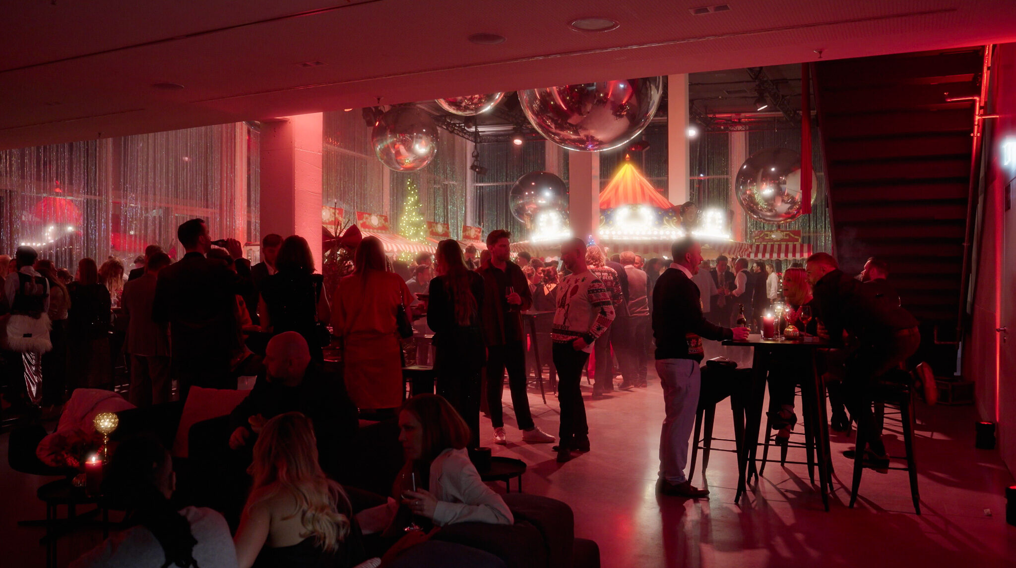 Eine stimmungsvolle, rot beleuchtete Weihnachtsfeier in einer modernen Eventlocation (Werk 3, München). Zahlreiche Gäste unterhalten sich in Gruppen an Stehtischen und auf Loungemöbeln. Die Decke ist mit großen, spiegelnden Silberkugeln dekoriert, während im Hintergrund ein beleuchtetes Karussell und Marktstände einen festlichen Jahrmarkt-Look erzeugen.