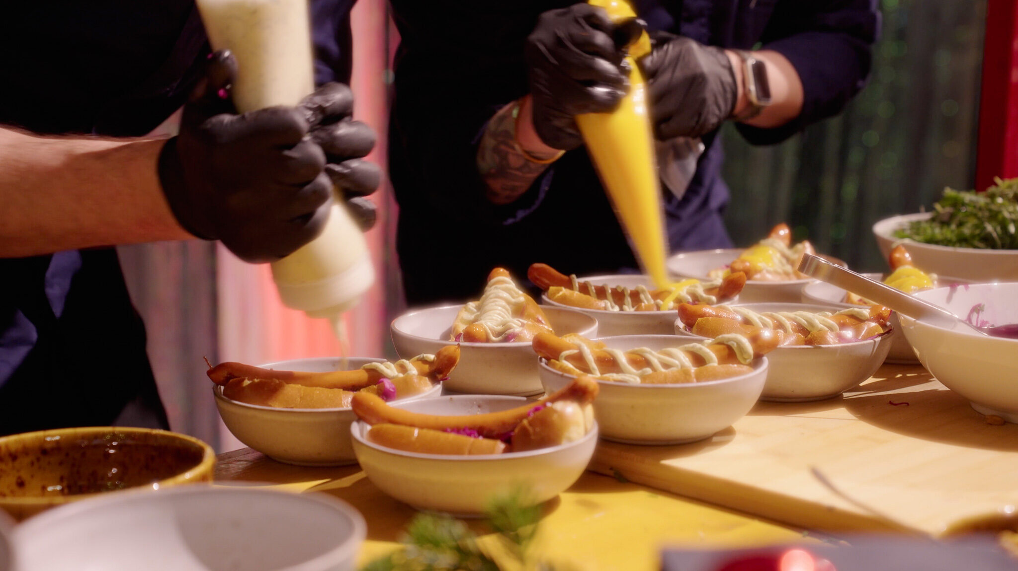Detailaufnahme vom Event-Catering bei der Avantgarde Weihnachtsfeier: Köche mit schwarzen Handschuhen bereiten frische Hot Dogs in Schalen zu und garnieren sie mit Saucen. Hochwertige Food-Fotografie von 4kFactory in München.