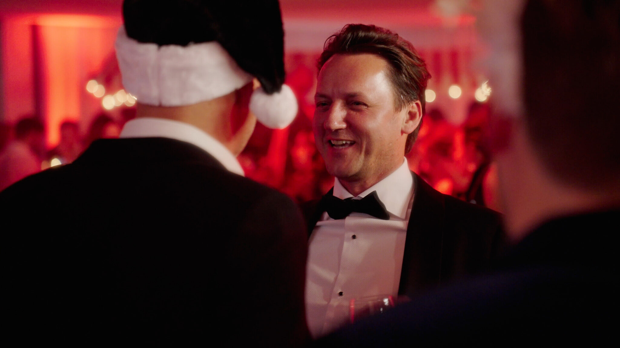 Ein emotionaler Moment auf der Avantgarde Weihnachtsfeier in München: Der neue CEO im Smoking lächelt im Gespräch mit dem ehemaligen CEO, der eine Weihnachtsmütze trägt. Close-up Event-Fotografie von 4kFactory in stimmungsvollem rotem Licht.