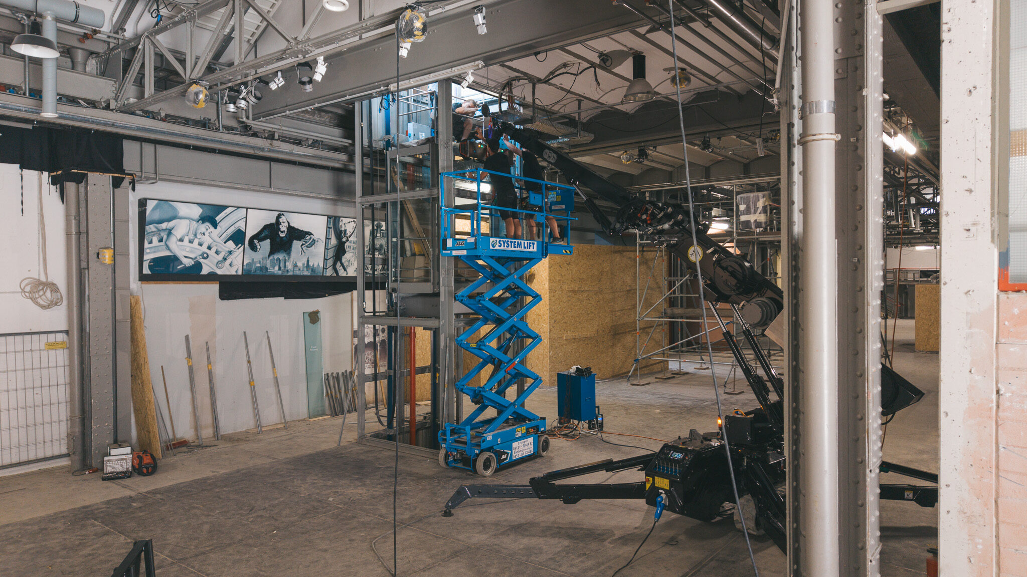 `alt="Innenansicht einer Baustelle in einer großen Industriehalle. Im Vordergrund liegen Baumaterialien und Werkzeuge auf dem staubigen Boden. In der Mitte stehen eine blaue Scherenarbeitsbühne und ein schwarzer Teleskopstapler. Die Decke ist offen mit sichtbaren Rohren und Kabeln, und im Hintergrund sind unfertige Wände und Gerüste zu sehen."`