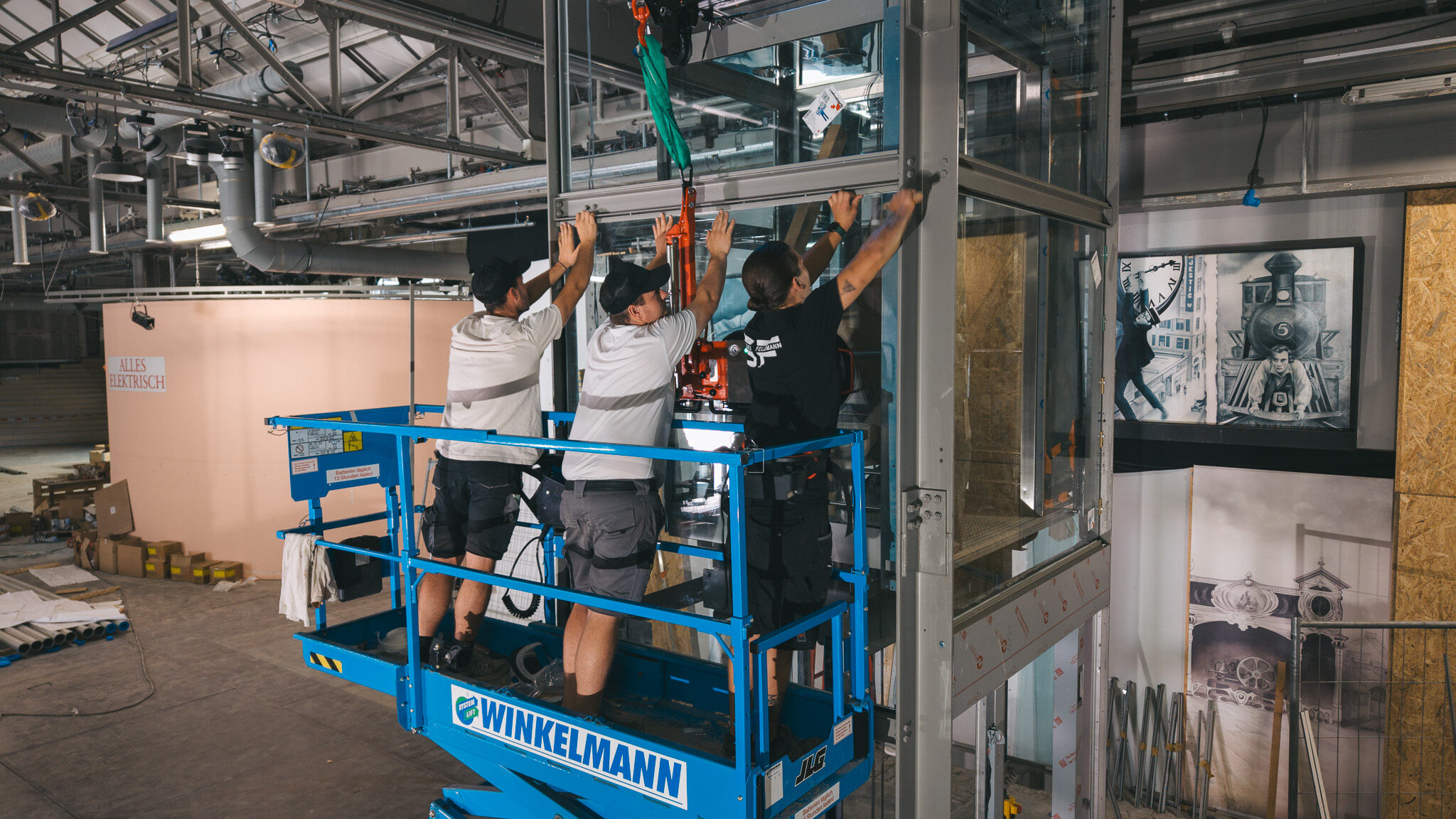 alt="Drei Modelle der 4kFactory arbeiten auf einer blauen Scherenbühne an einer Glasinstallation in einer Industriehalle. Im Hintergrund ist ein künstlerisches Wandbild mit Lokomotiv-Motiv zu sehen. Das ist ein Werbefilm für die Glaserei Feldmann"