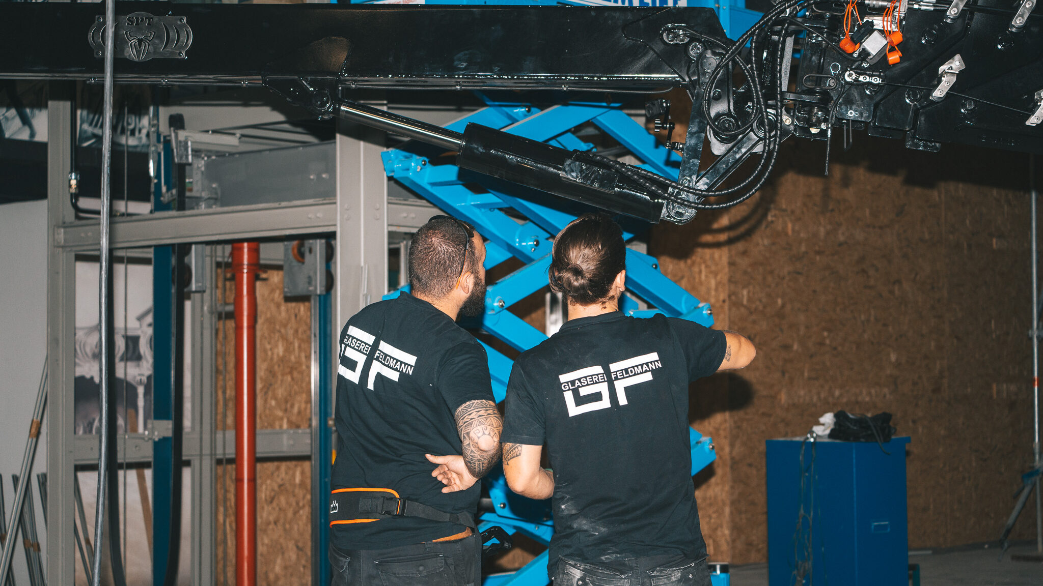 alt="Baustellen-Dokumentation der 4kFactory für Glaserei Feldmann. Spezialisten montieren eine komplexe Glas-Konstruktion für einen Aufzugsschacht in einer modernen Werkhalle."