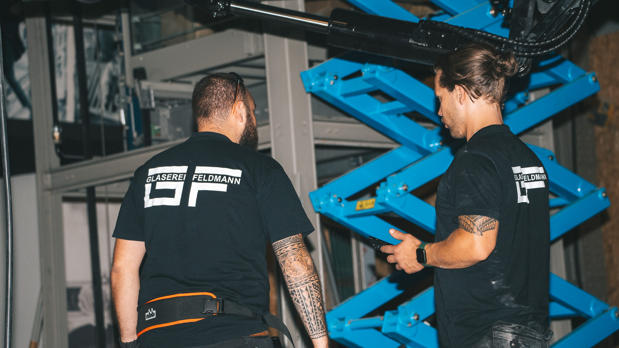 alt="Baustellen-Dokumentation der 4kFactory für Glaserei Feldmann. Spezialisten montieren eine komplexe Glas-Konstruktion für einen Aufzugsschacht in einer modernen Werkhalle."