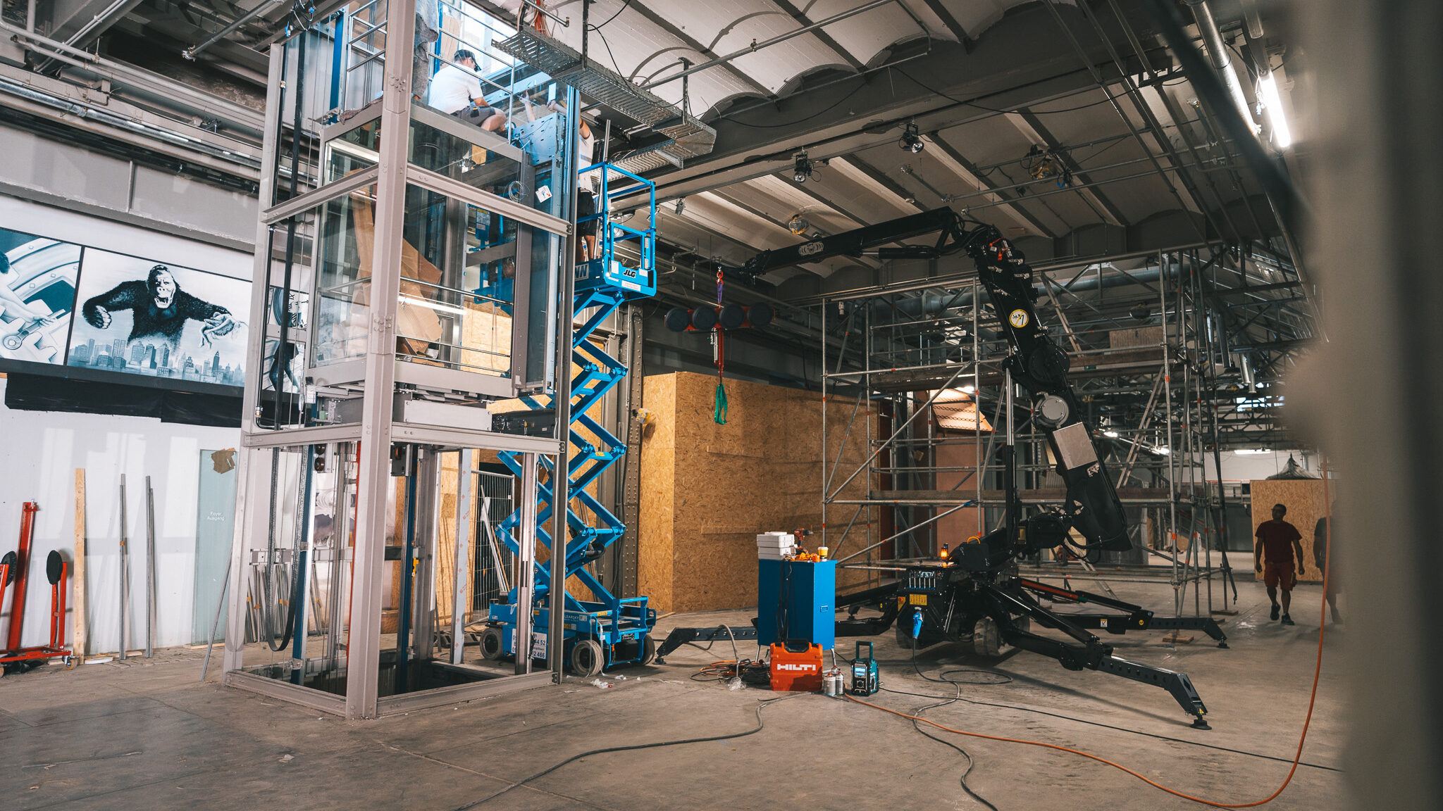 alt="Mitarbeiter der Glaserei Feldmann bei Montage-Arbeiten auf einer blauen Scherenbühne. Dokumentiert von 4kFactory in einer Industriehalle in Nürnberg beim Schulmuseum."