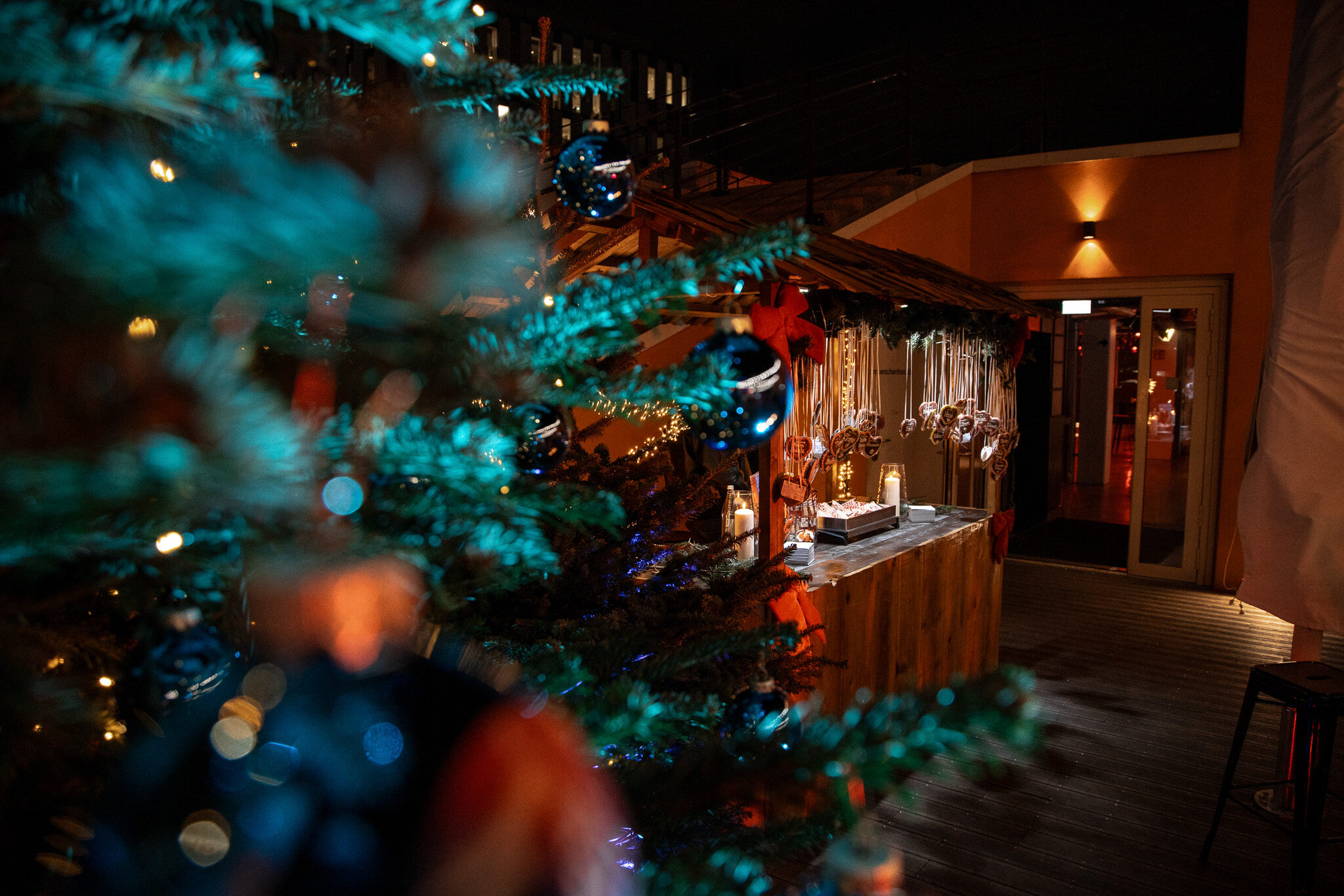 Stimmungsvolle Eventfotografie mit Bokeh-Effekt: Weihnachtsbaum im Vordergrund und beleuchteter Marktstand bei der Avantgarde Firmenfeier, umgesetzt von 4kFactory für das event video München.