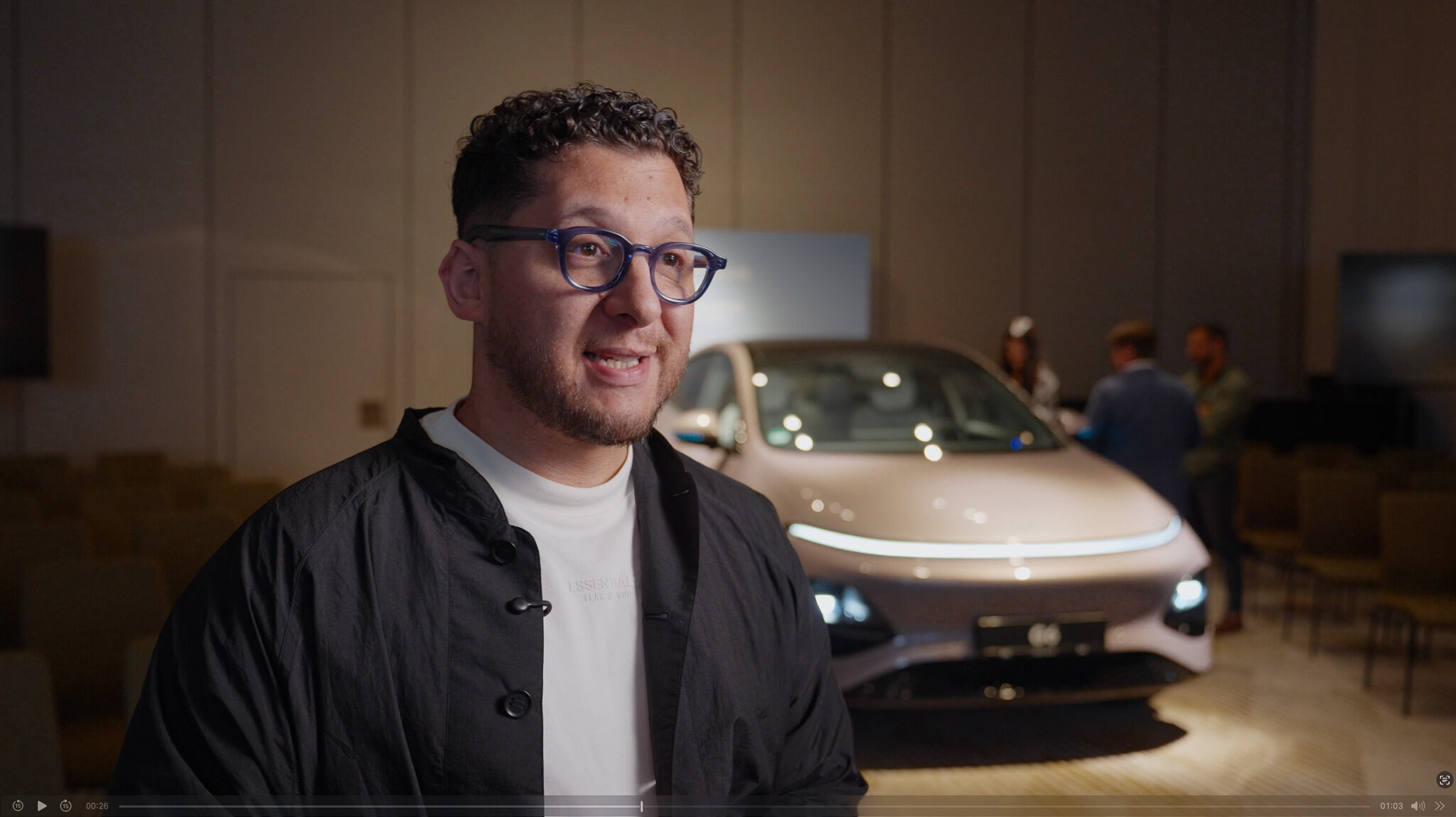 „Ein Mann mit Brille und dunkler Jacke in einer Interviewsituation während einer Automotive-Videoproduktion, im Hintergrund ist ein XPENG G6 Elektroauto dezent beleuchtet.“