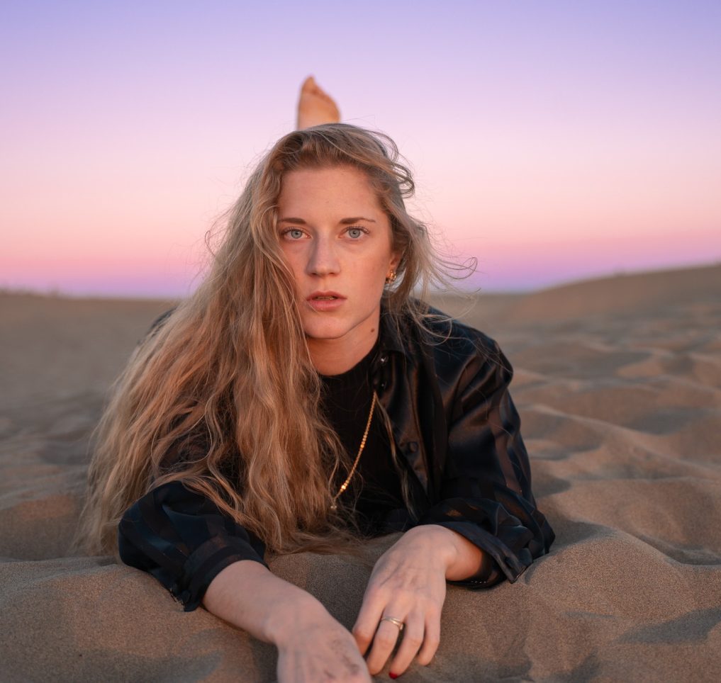 Eine Frau liegt auf Sand. Hinter ihr sind Dünen zu sehen. Es ist Sonnenuntergang.