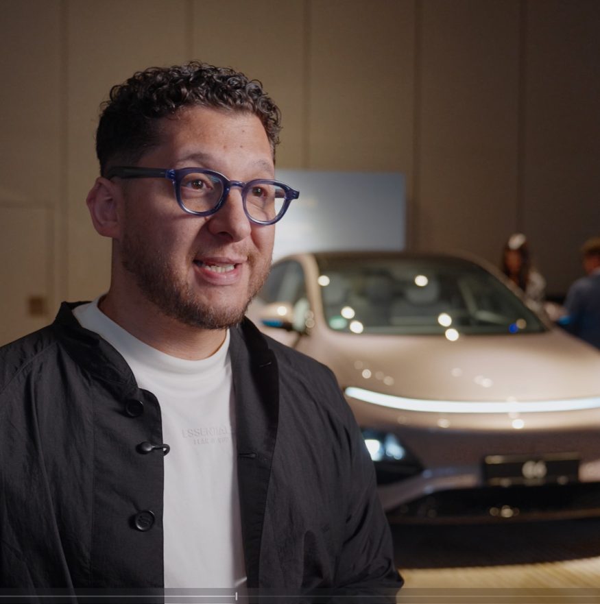 „Ein Mann mit Brille und dunkler Jacke in einer Interviewsituation während einer Automotive-Videoproduktion, im Hintergrund ist ein XPENG G6 Elektroauto dezent beleuchtet.“